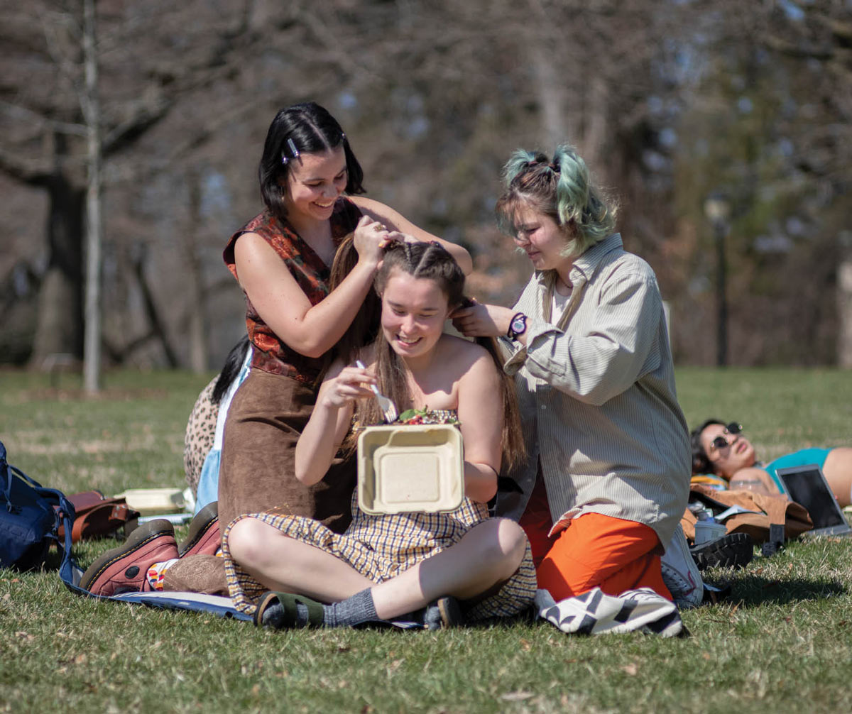 two Swarthmore students braiding the hair of another Swarthmore student and they eat lunch on the grass