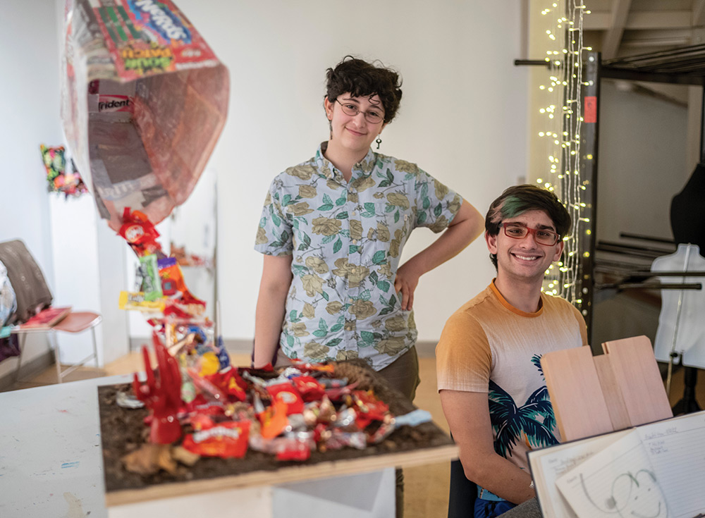 Two Swarthmore College students smile and pose as one of them is standing and the other is sitting while both of them are nearby a table of candy