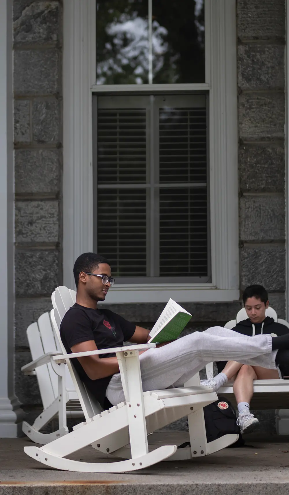 Two Swarthmore College students reading their own books as one of the guys has see through prescription glasses and has his legs extended out on a white wooden rocking chair while the other guy is sitting on a non-rocking chair