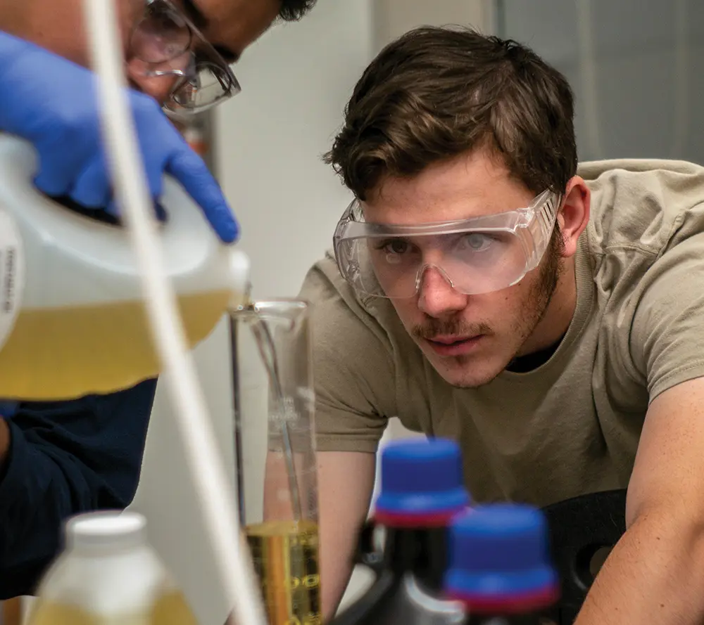 Two Swarthmore College students wearing safety goggles as they examine liquid pouring into a thin/skinny chrome colored beaker as one of the guys is holding a jug full of liquid to pour into the object