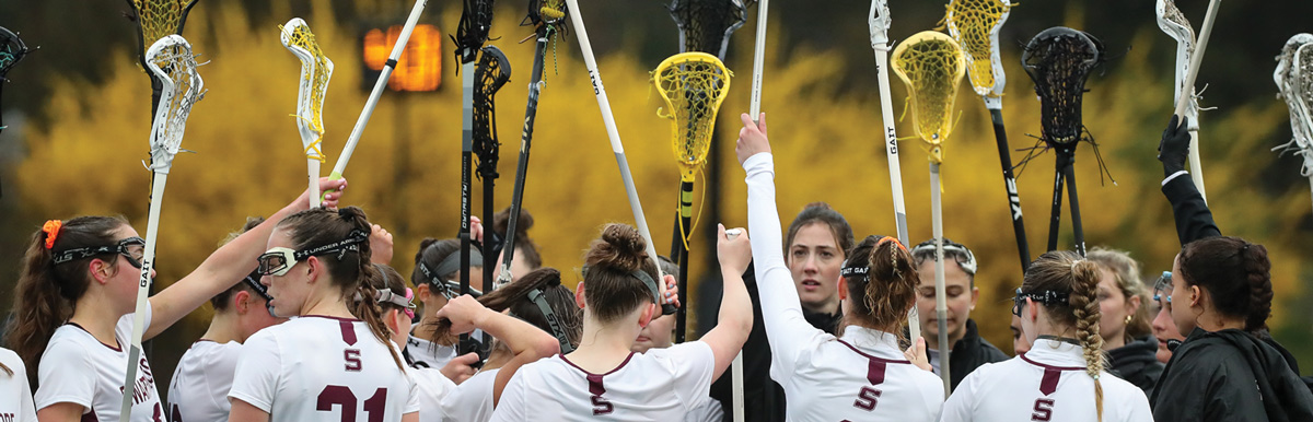 Lacrosse team in a huddle with sticks raised, wearing white jerseys with maroon details against a backdrop of yellow foliage.