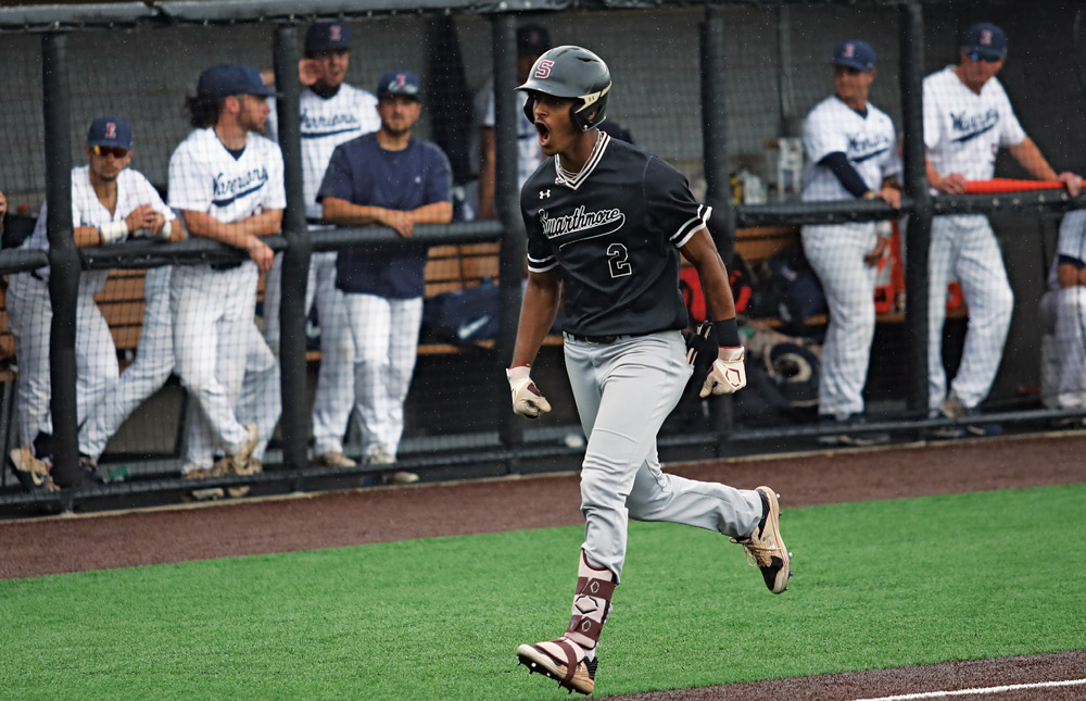 A baseball player in a black uniform runs past a dugout with a team in white uniforms watching.