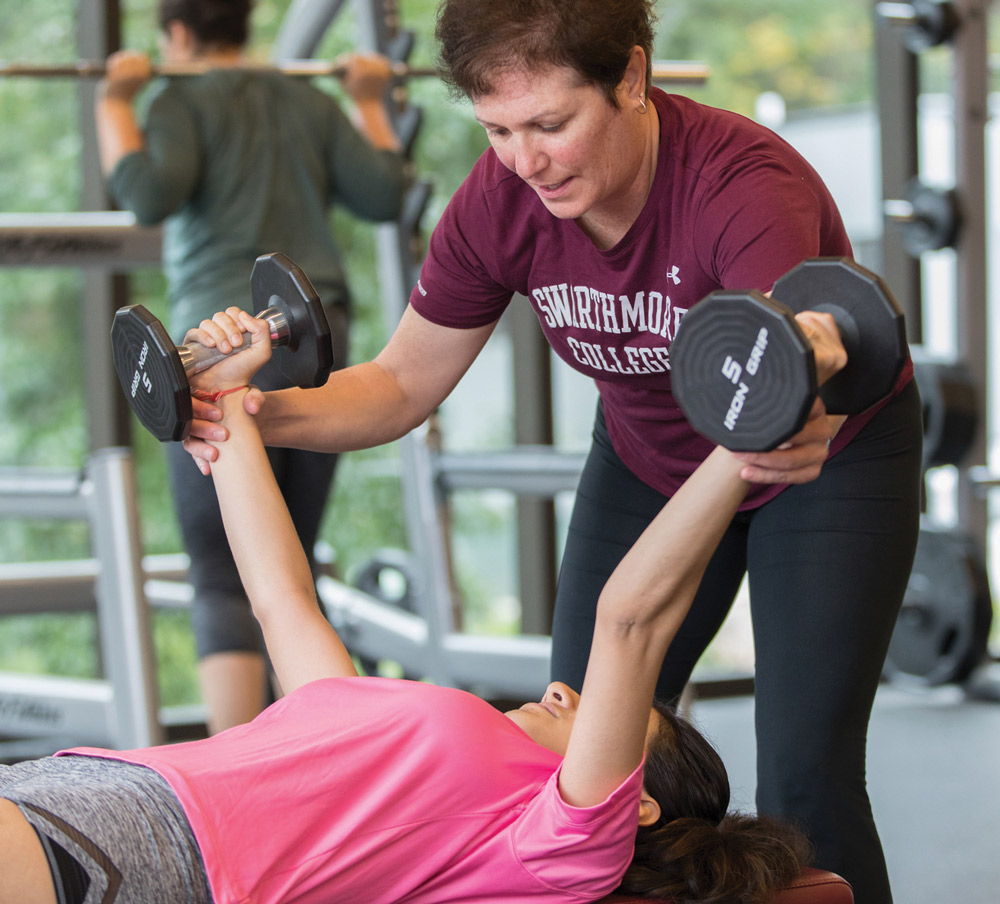 A gym instructor in a maroon shirt assists someone lifting dumbbells on a bench.