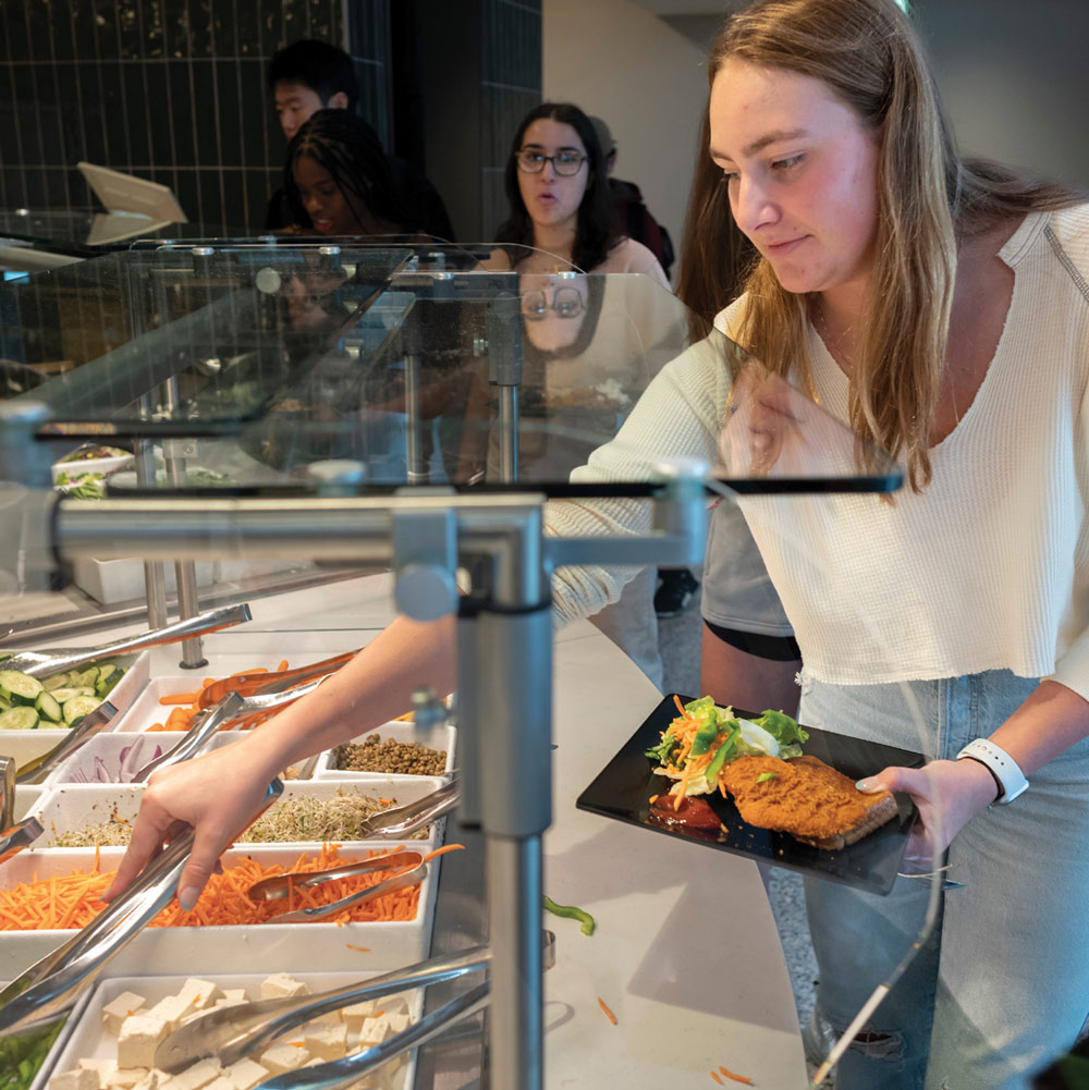 A woman at a salad bar selecting food with tongs while holding a black plate.