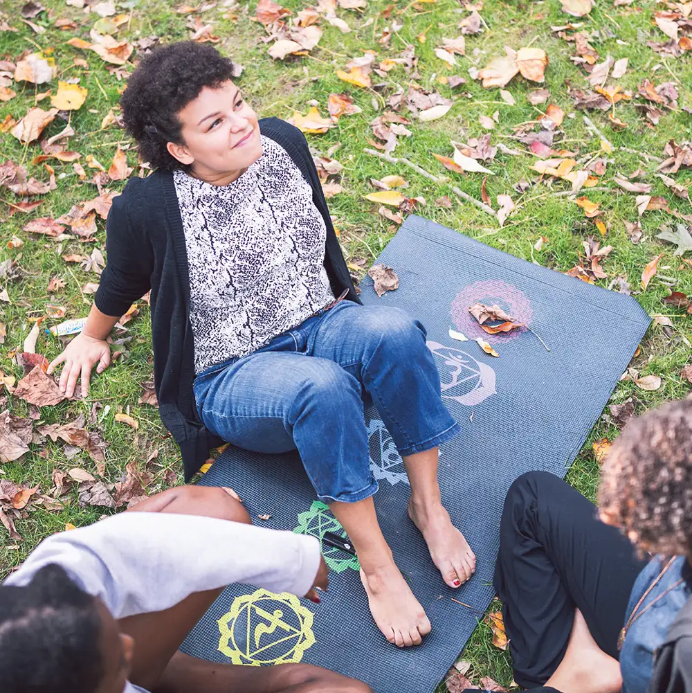Person sitting on a yoga mat adorned with chakra symbols on a grassy lawn with autumn leaves.