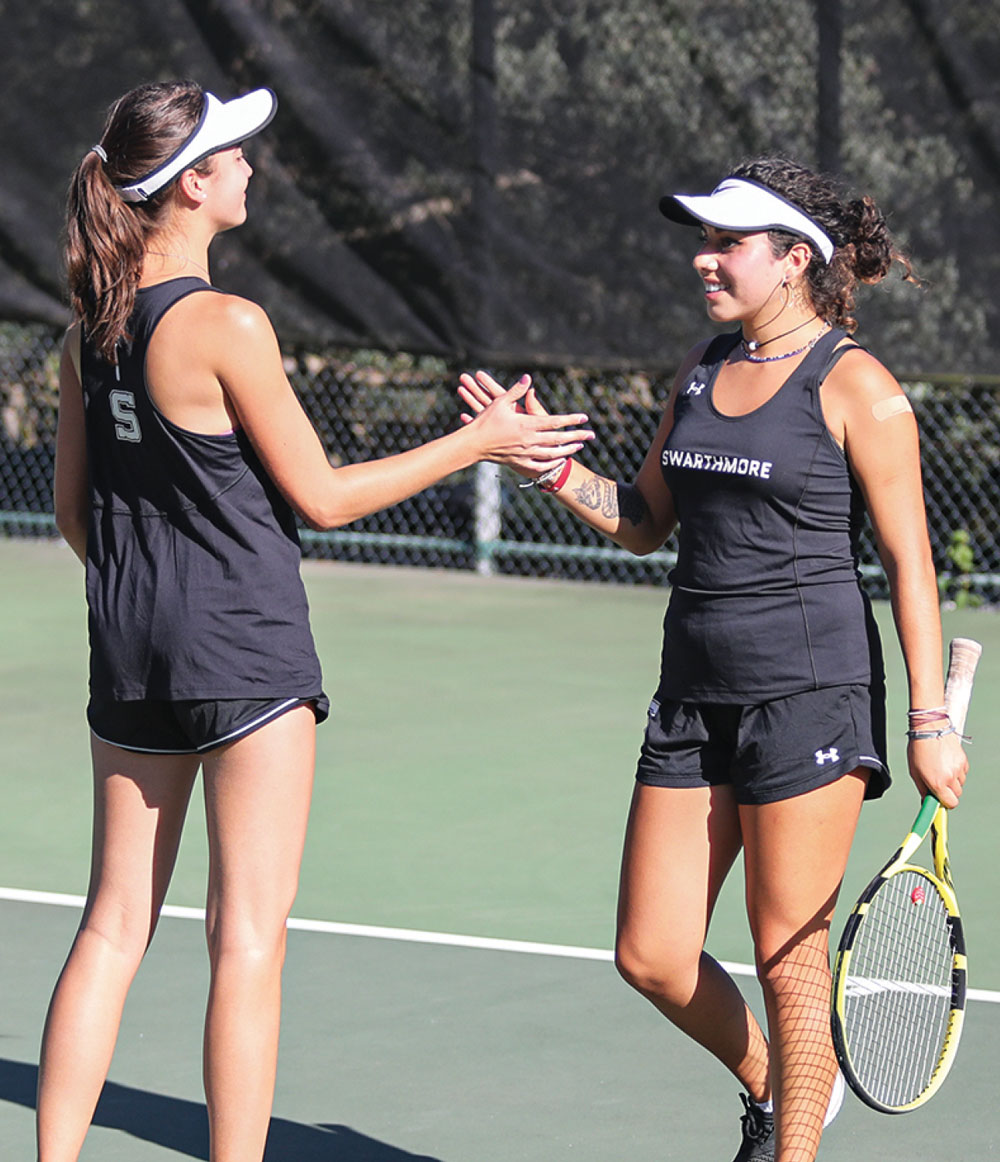 Two female tennis players in black outfits and white visors shaking hands on a court.