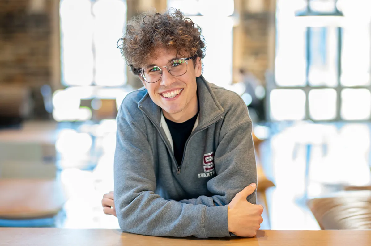 Portrait of a young man smiling and leaning on a table indoors. He wears glasses and a gray Swarthmore sweatshirt.