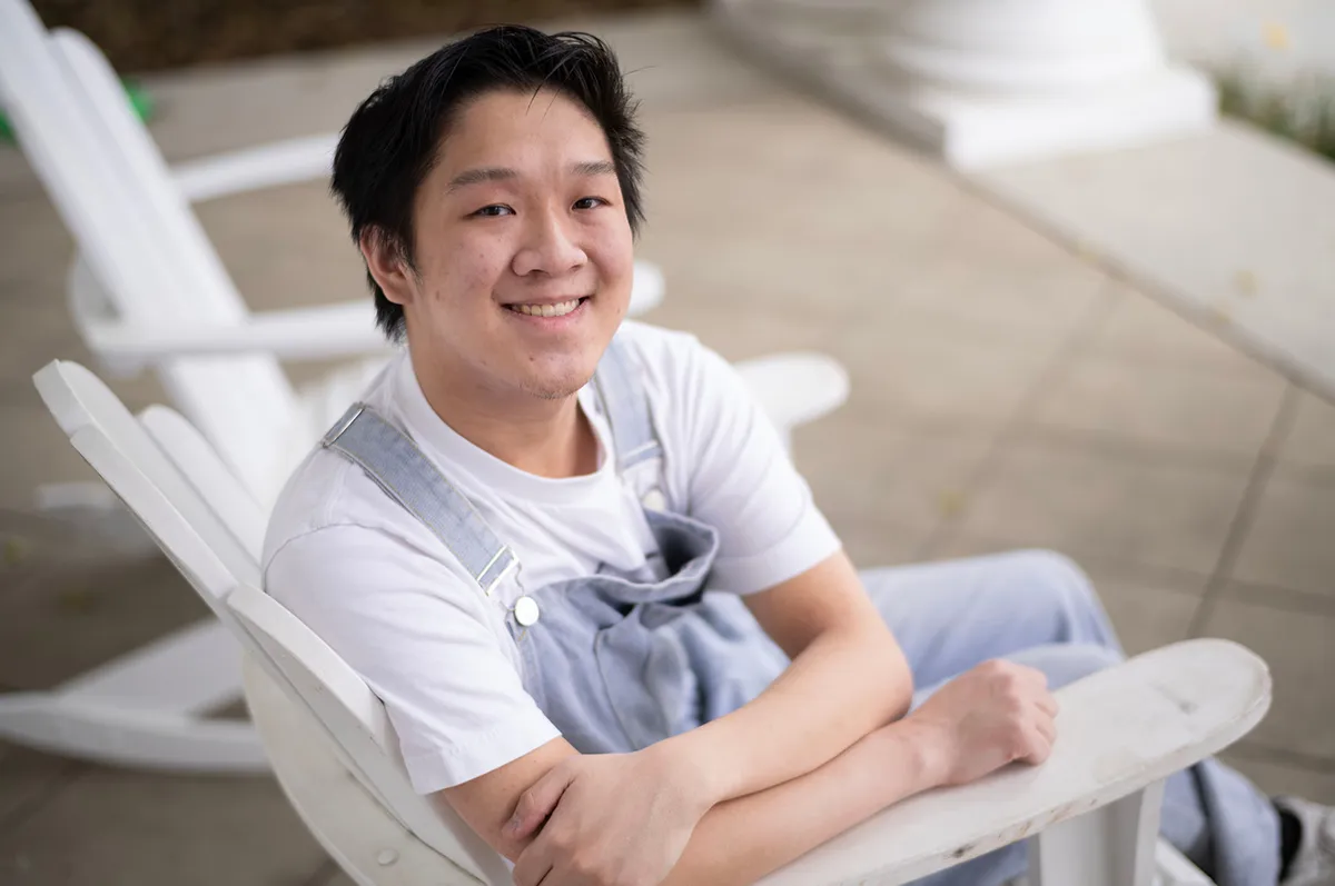 Portrait of a young man seated in a white outdoor chair, smiling. He wears denim overalls over a white shirt.