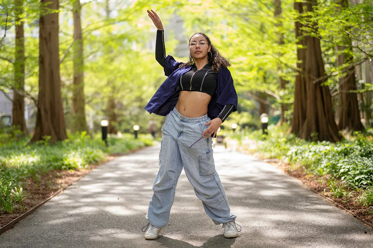 Portrait of a young woman striking a confident dance pose on a tree-lined path. She wears a black crop top, baggy jeans, and a purple shirt.