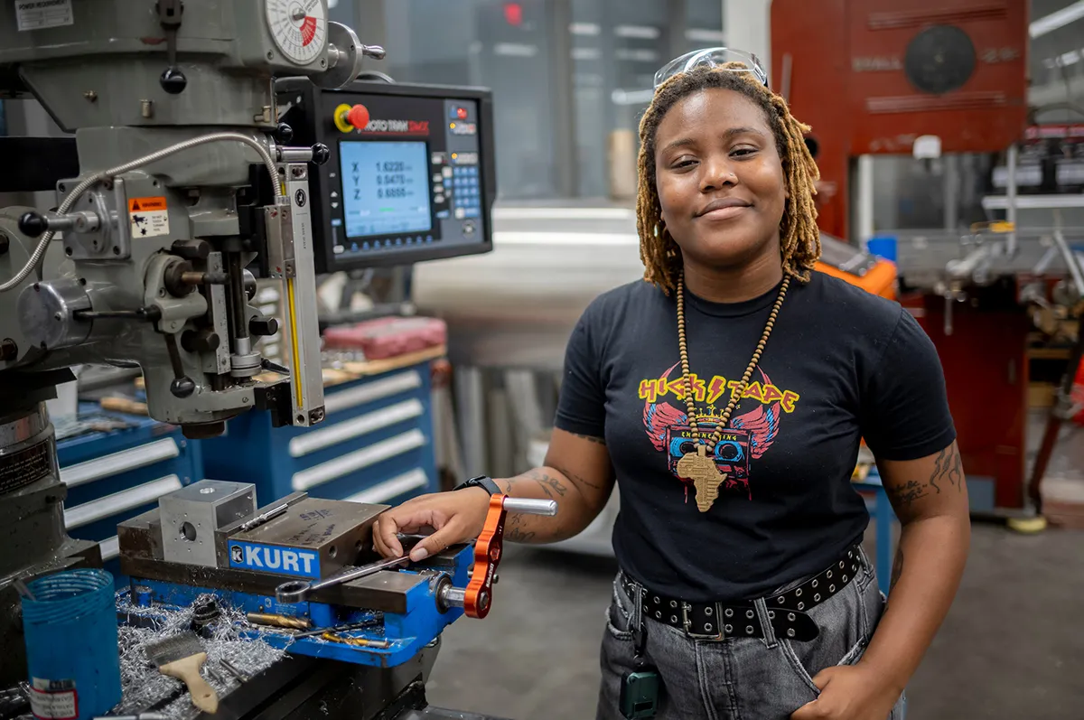 Portrait of a young woman standing in a machine shop, smiling confidently. She wears a black graphic T-shirt, jeans, and protective glasses on her head.