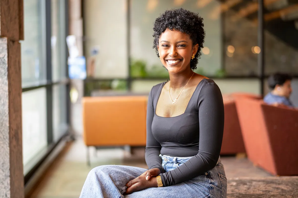 Portrait of a young woman sitting indoors on a bench, smiling warmly. She wears a black top, jeans, and gold accessories.