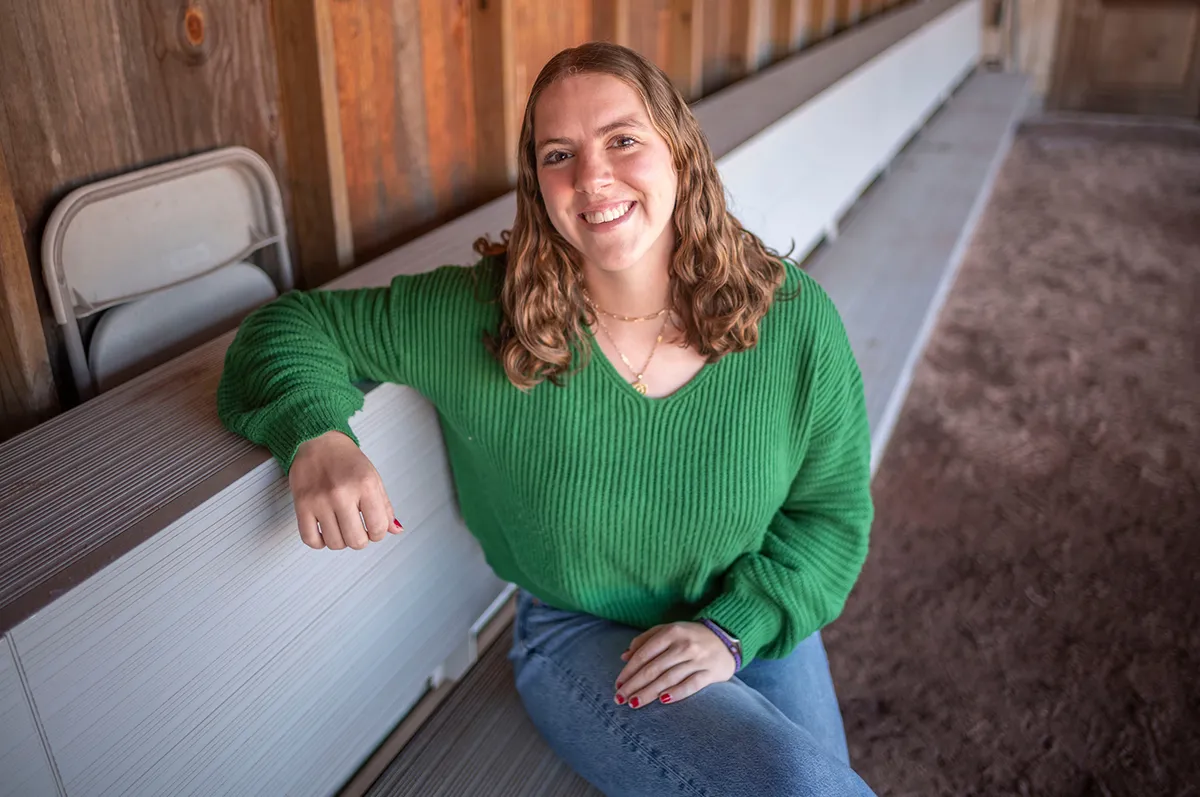 Portrait of a young woman sitting on bleachers in a wooden building. She wears a bright green sweater and jeans and is smiling.