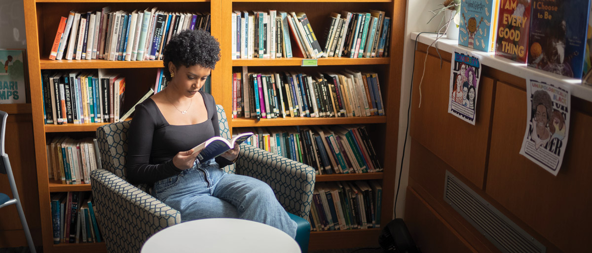 A person sits in a library reading a book, surrounded by shelves filled with books.