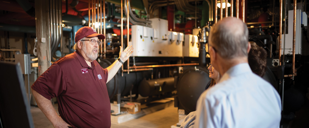 A man in a maroon shirt gestures while speaking in an industrial room with pipes and machinery.