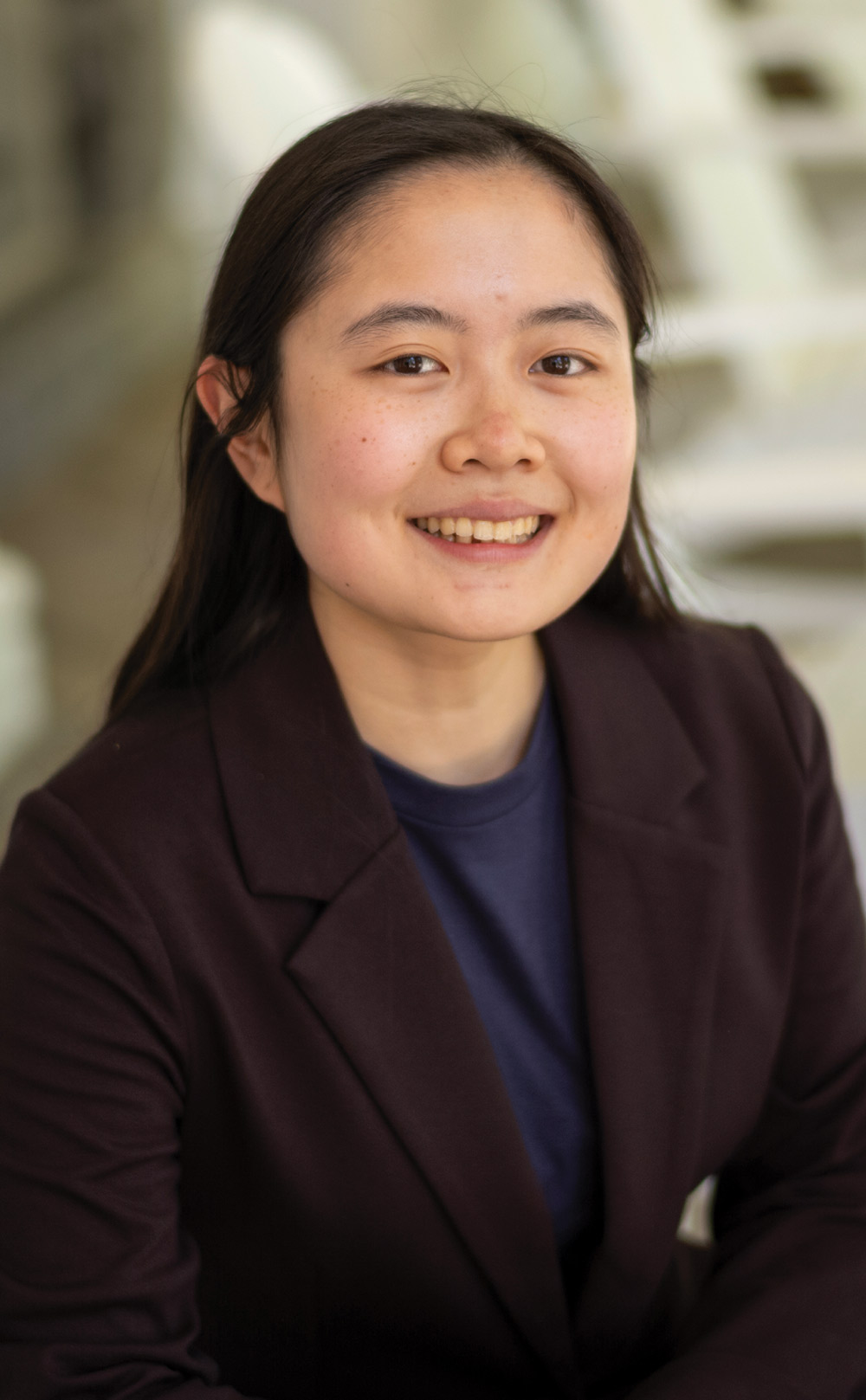 Close-up portrait of a smiling young Asian woman with long dark hair, wearing a dark blazer over a blue shirt.