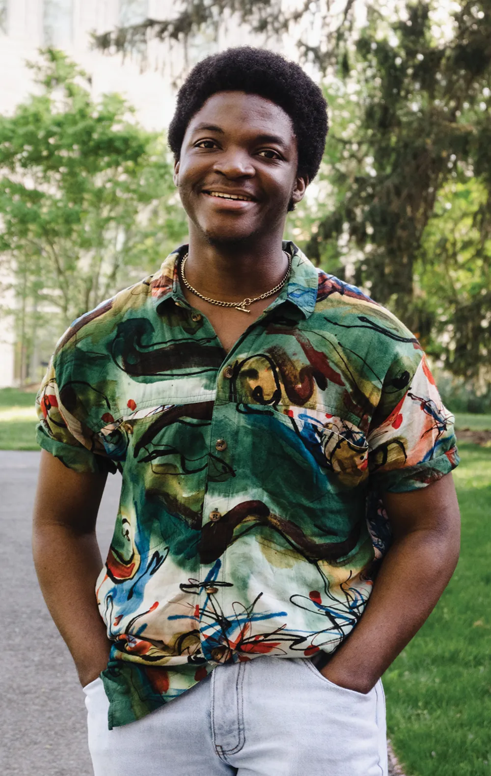 A smiling young Black man with an afro stands outdoors, wearing a colorful abstract-pattern button-up shirt and a gold chain.