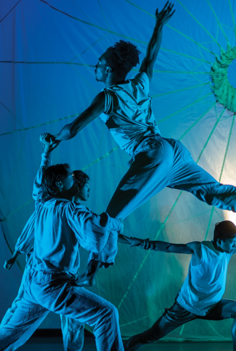 Dancers perform a complex lift under vibrant blue and green stage lights, showcasing athletic, mid-air choreography.