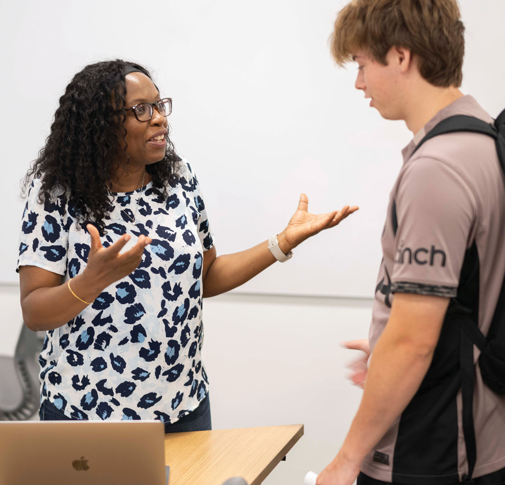 A Black woman professor gestures while talking to a young male student in a classroom setting next to a laptop.
