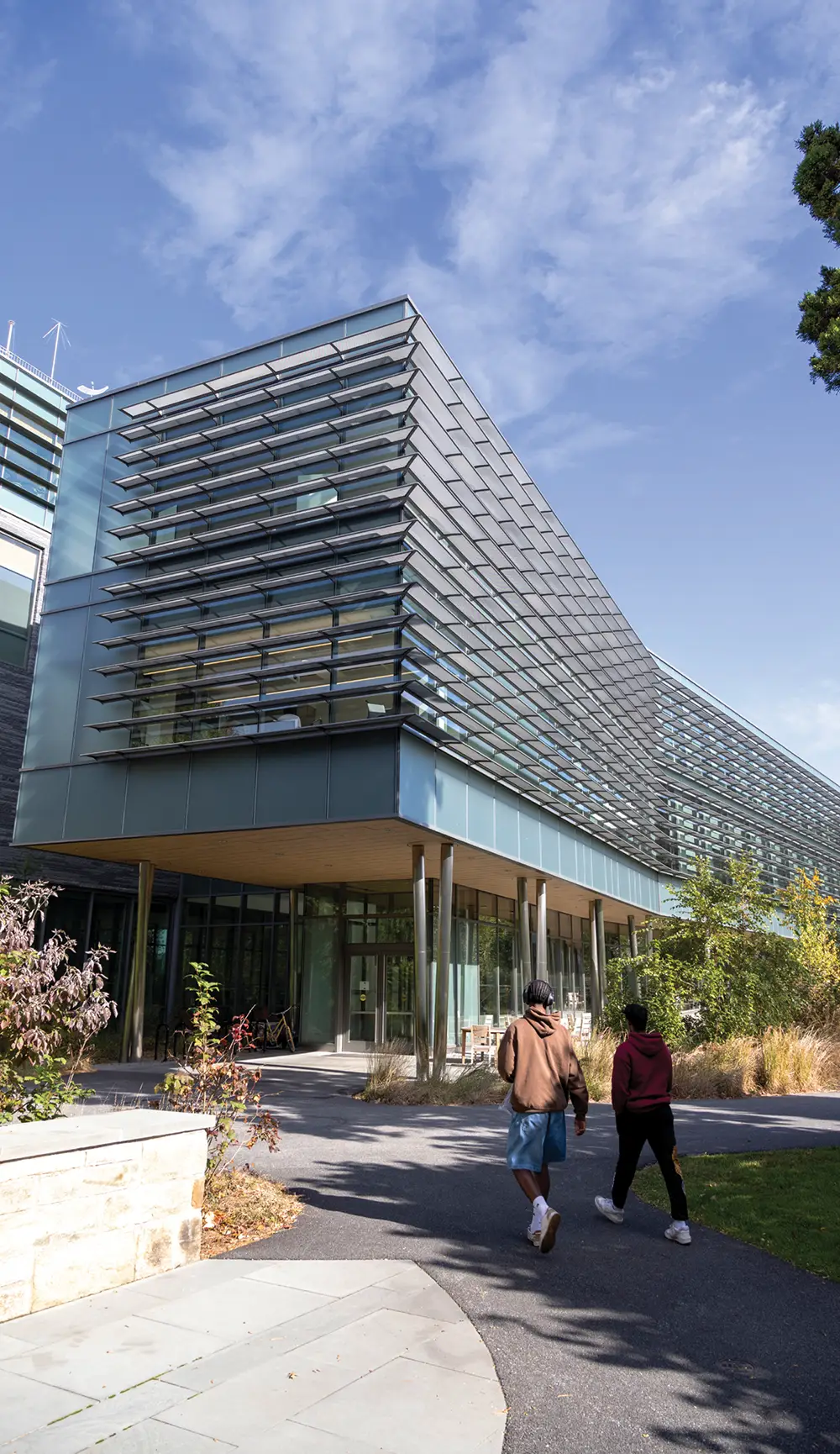 Two students walk toward a modern glass and steel campus building with large horizontal window louvers on a sunny day.