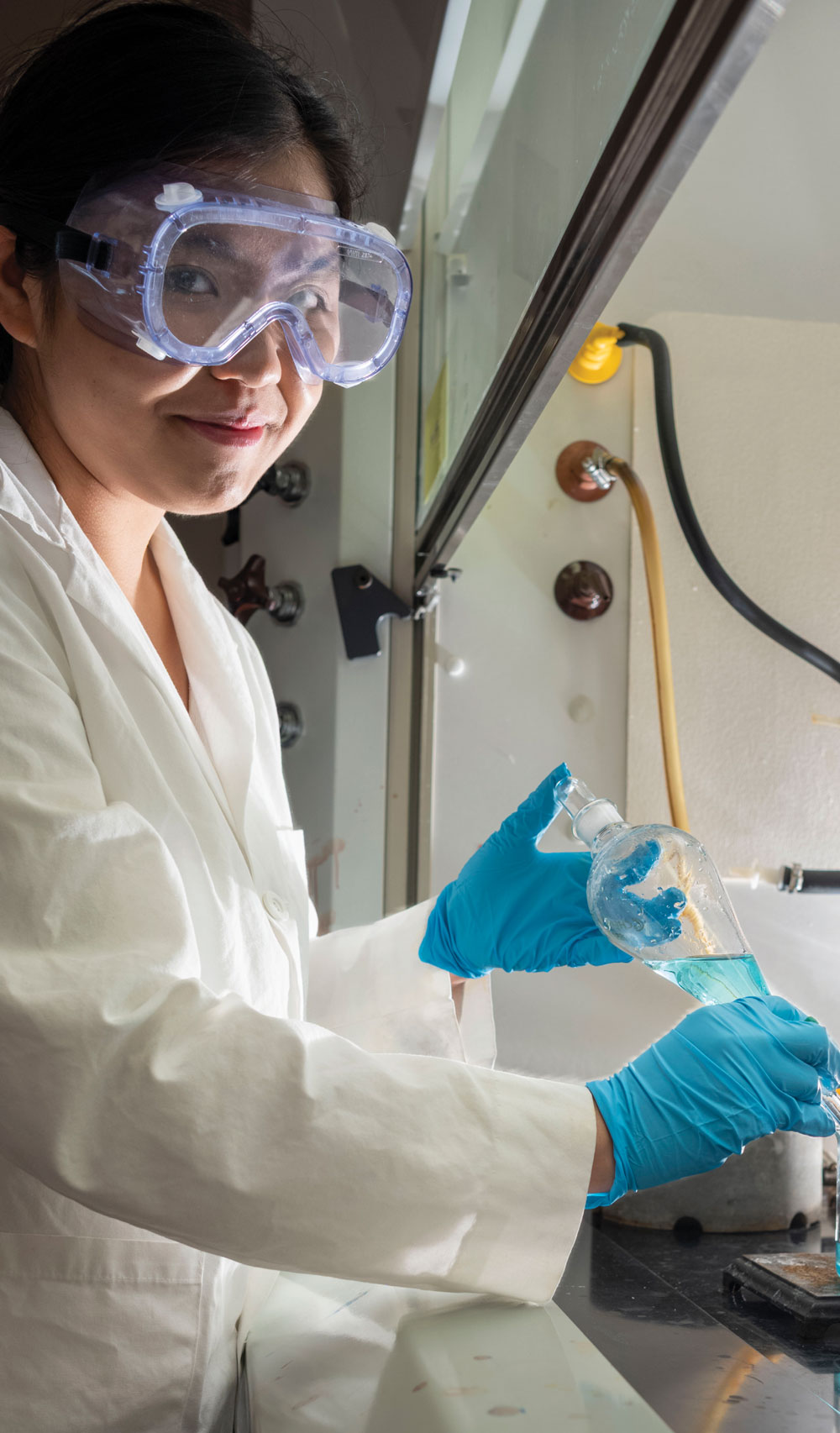 A smiling student in a lab coat and safety goggles handles a glass flask containing blue liquid inside a fume hood.