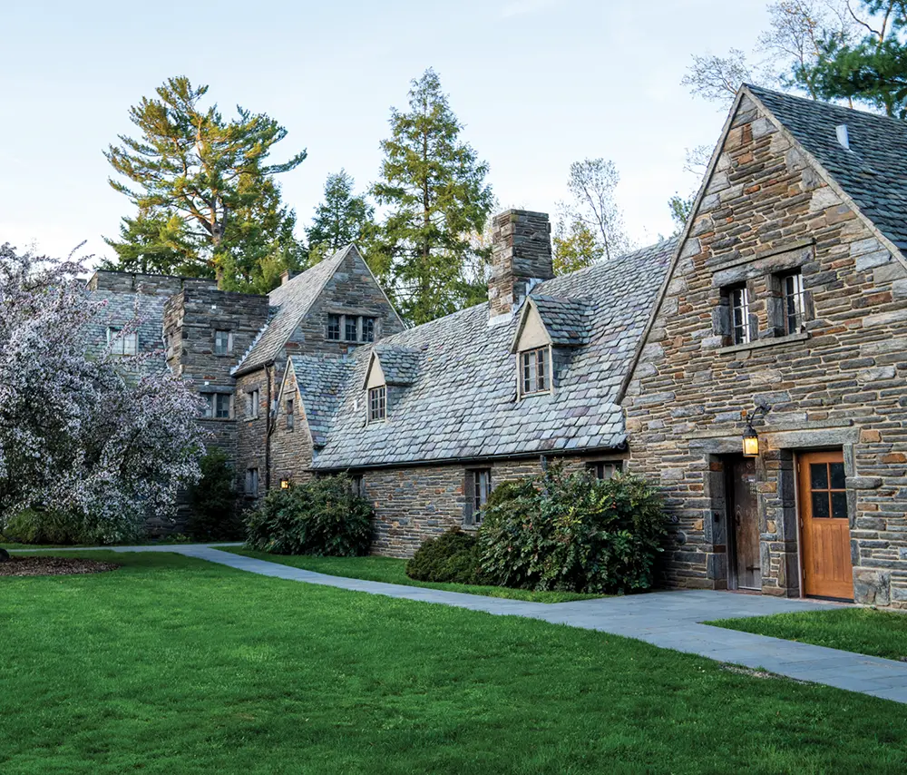 Exterior of a historic, multi-story stone residence hall with a slate roof, surrounded by green lawns and trees.