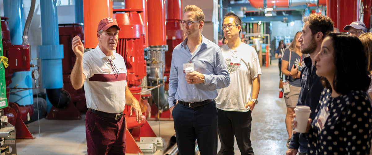 A guide in a red cap gestures as he leads a tour through a campus utility facility with large red and blue pipes.