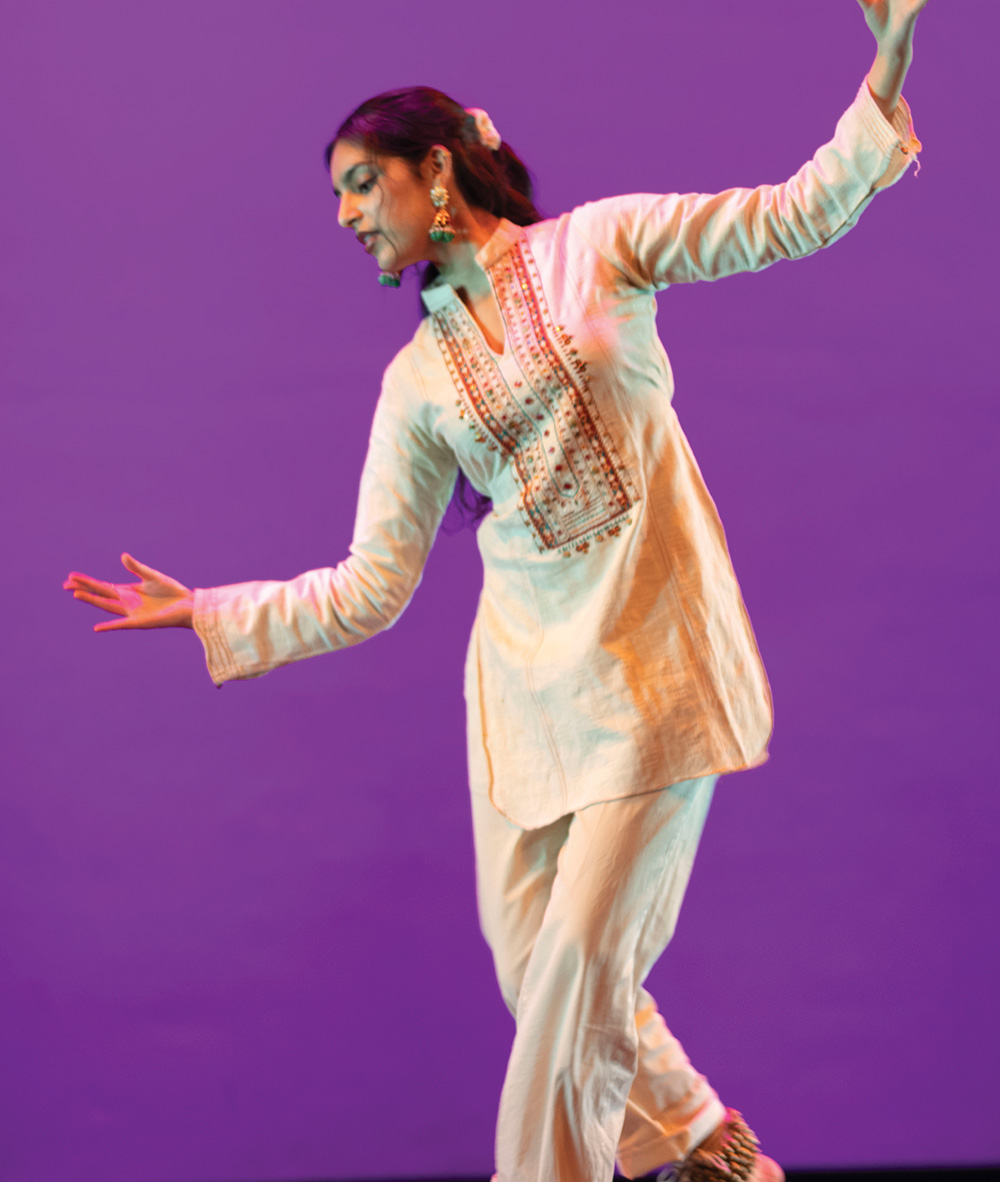 A student performs a traditional dance on stage in an embroidered cream tunic and trousers against a purple backdrop.