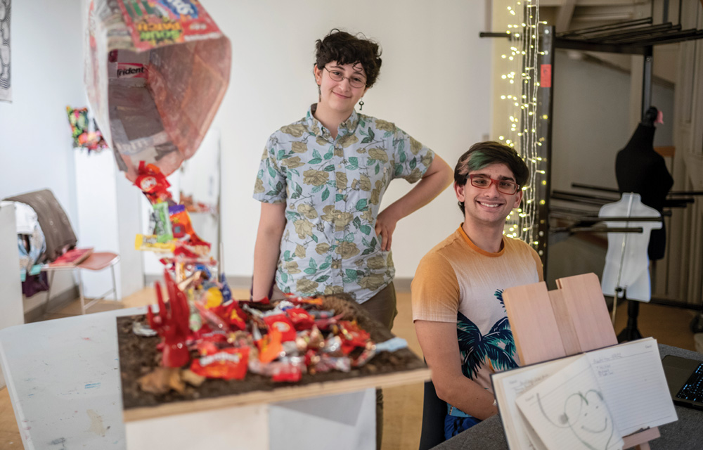 Two students smile near an art installation made of colorful candy wrappers and sculpture materials in a studio.