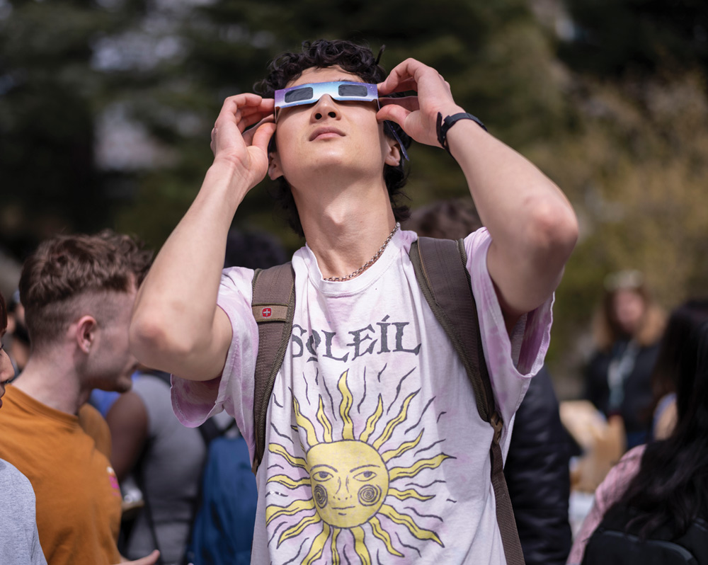 A student wearing protective solar eclipse glasses looks up at the sky during an outdoor gathering on campus.