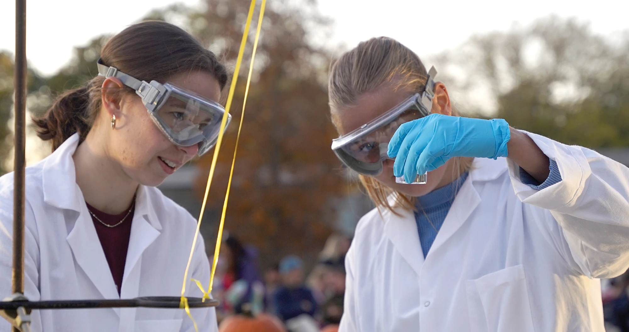 Two students wearing lab coats and safety goggles closely observe a liquid solution in a beaker outdoors.