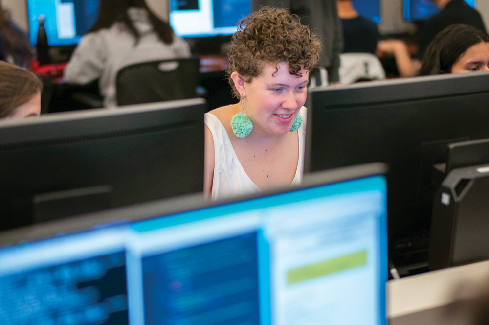 A smiling student wearing green pom-pom earrings works at a computer screen in a busy computer lab.