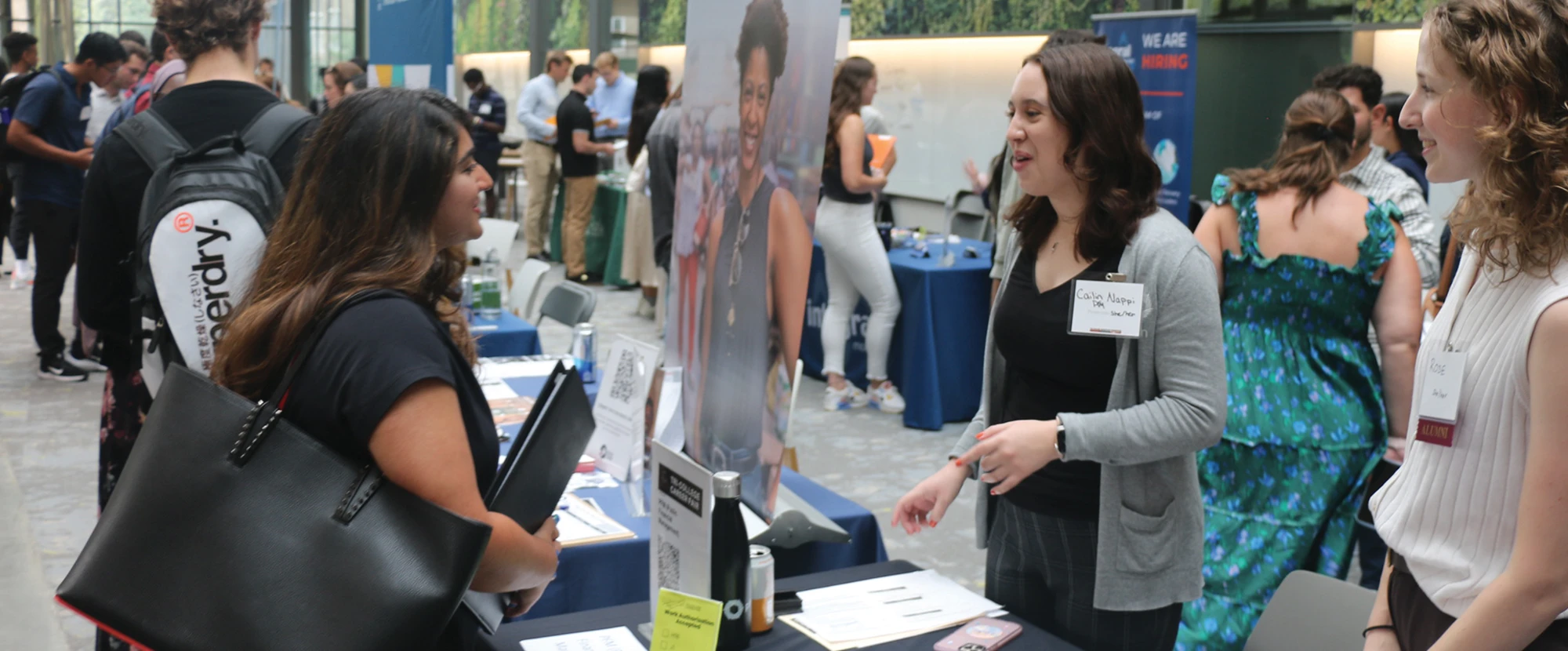 A student holding a binder speaks with a representative at a recruitment booth during a crowded campus career fair.