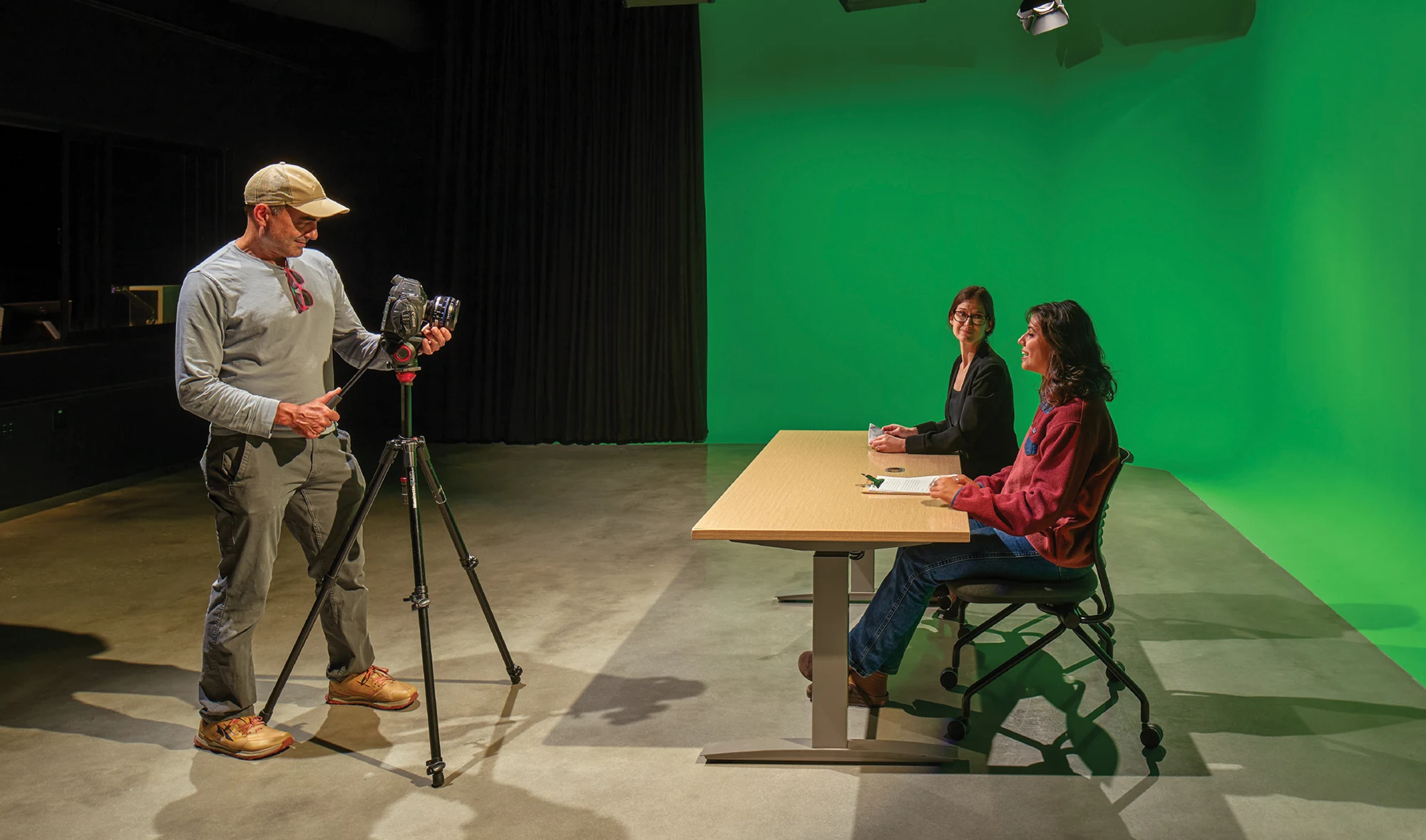 A videographer films two women seated at a desk in front of a large green screen inside a media production studio.