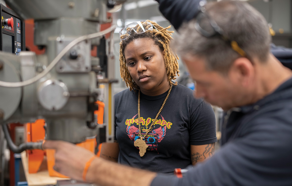 An engineering student wearing safety glasses learns to operate machinery under instructor guidance in a lab.