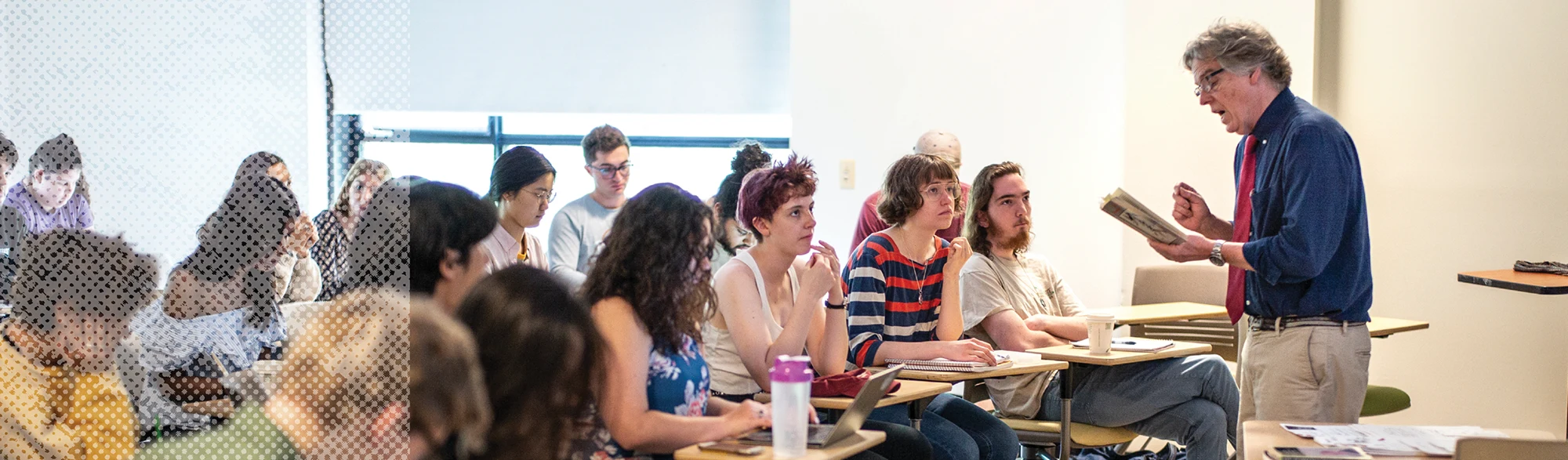 A professor holding a book lectures in front of attentive students seated at desks in a bright classroom.