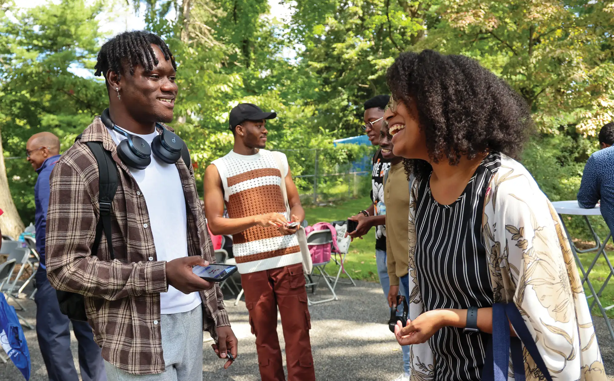 A group of students laugh and converse outside during a sunny campus event surrounded by lush green trees.