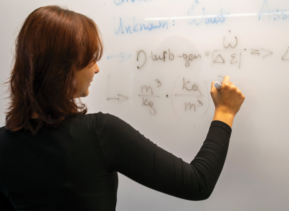 A student writes complex physics or engineering formulas on a white dry-erase board with a marker.