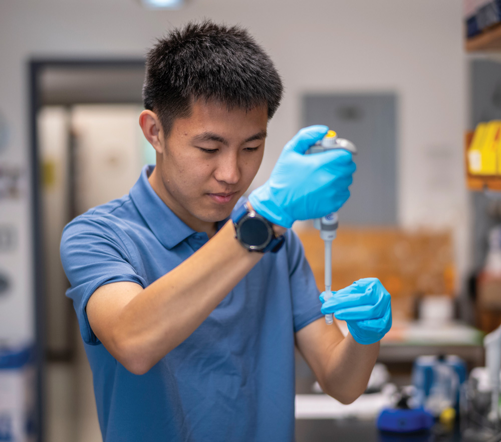 A student in a blue polo and gloves uses a micropipette to carefully transfer liquid into a vial in a science lab.