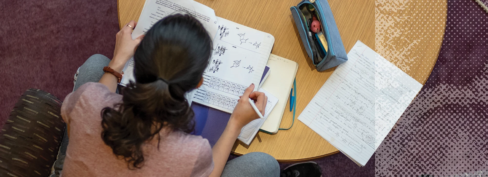Overhead view of a student studying chemistry notebooks and handwritten notes at a round wooden table.