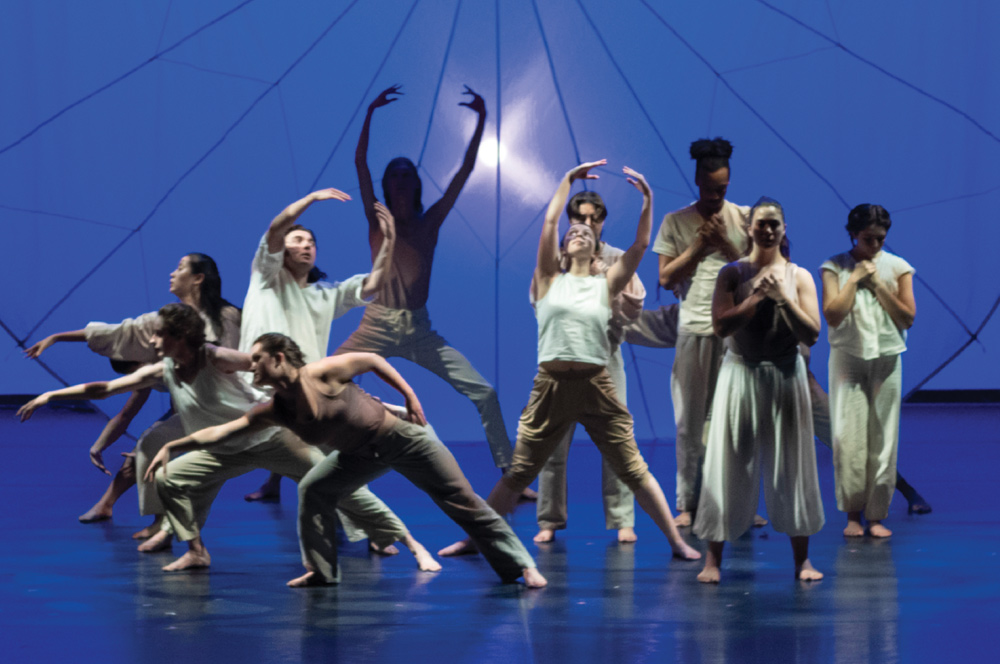 A group of dancers in neutral clothing perform expressive choreography together on stage under blue spotlighting.