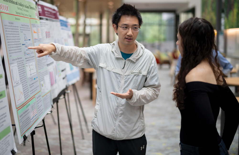 A student points to data on an academic poster display while explaining his research project to a peer.